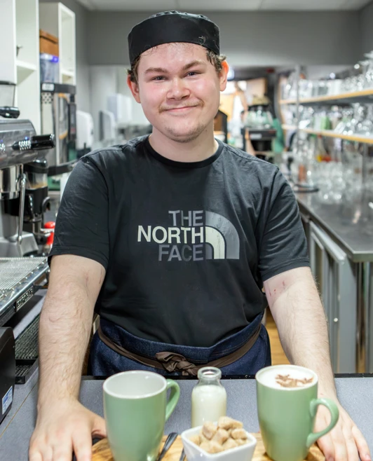 Barista student proudly presenting a tray with two cups of coffee, sugar cubes, and milk in a café setting. Barista student proudly presenting a tray with two cups of coffee, sugar cubes, and milk in a café setting.
